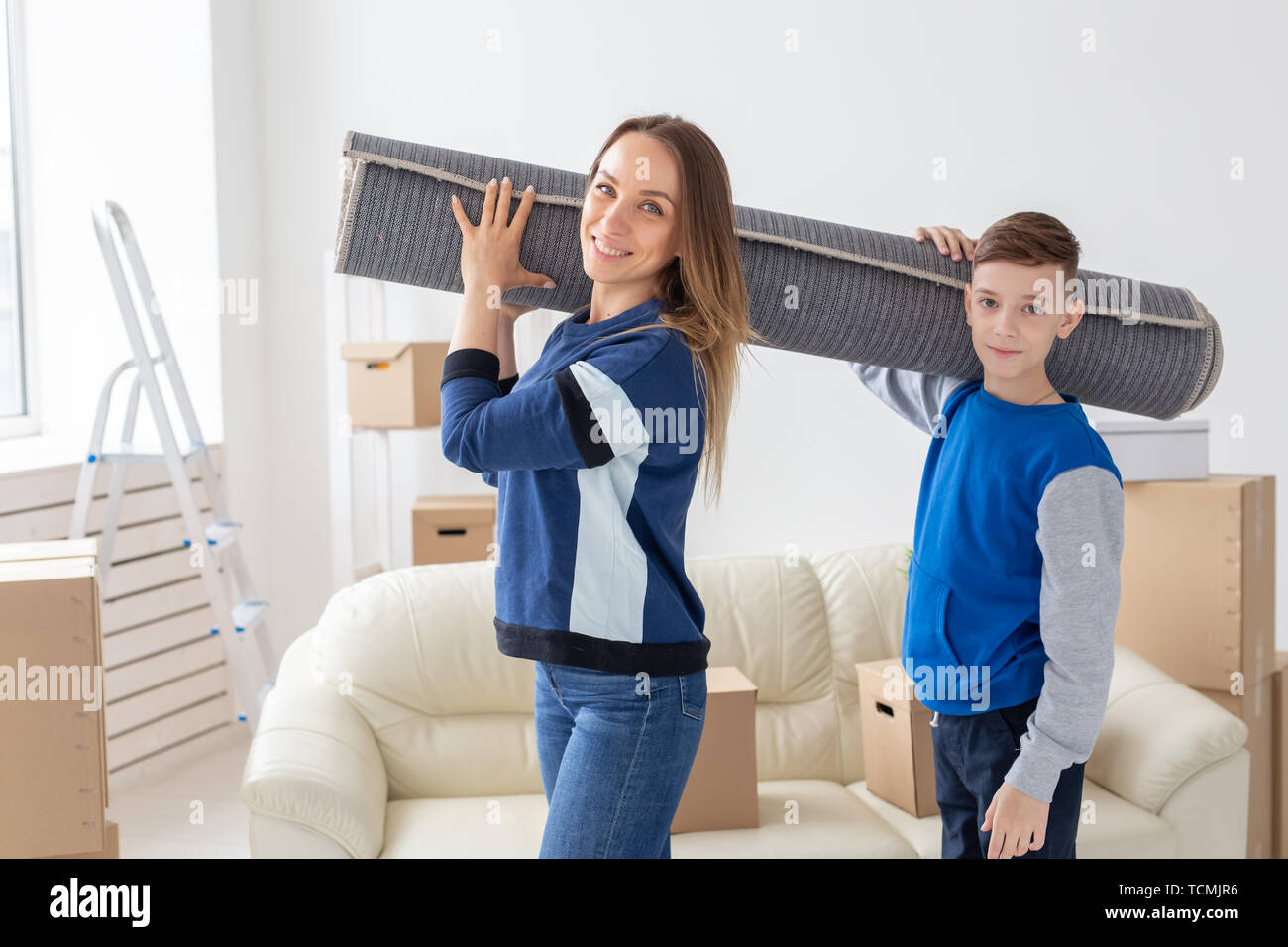 Smiling caucasian mother and a charming son are holding a folded carpet on their shoulders in a new living room intending to spread it in a new Stock Photo
