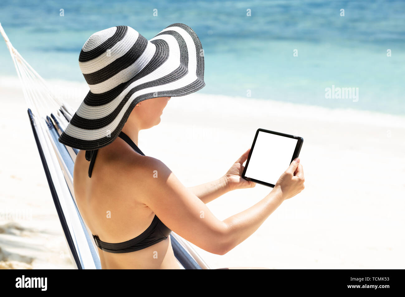 Rear View Of A Woman In Bikini Sitting On Hammock Using Digital Tablet At Beach Stock Photo