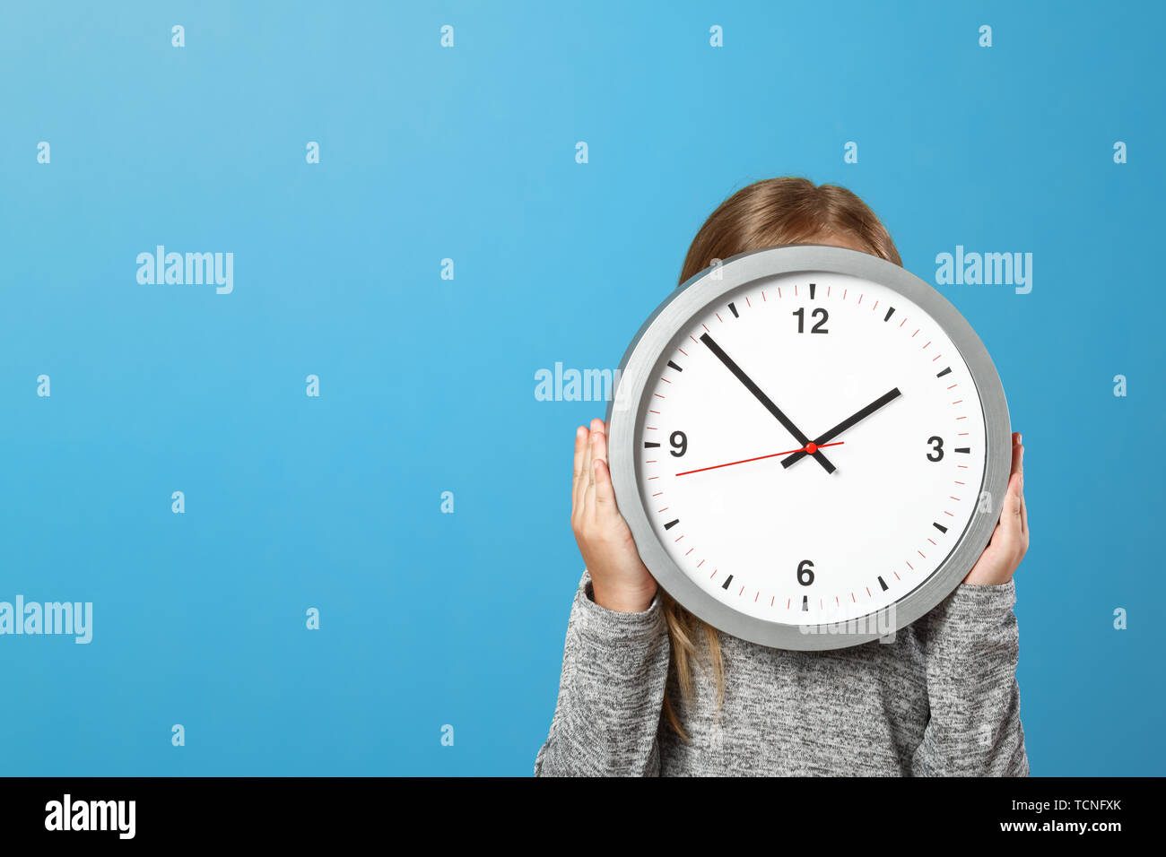 The little girl is hiding behind a big clock on a blue background. The concept of education, development, school, timing, time to learn. Stock Photo