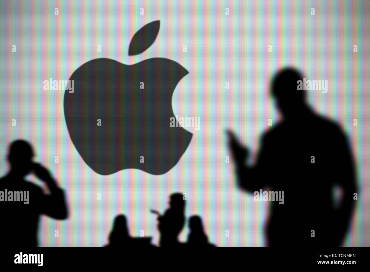The Apple logo is seen on an LED screen in the background while a silhouetted person uses a smartphone in the foreground (Editorial use only) Stock Photo