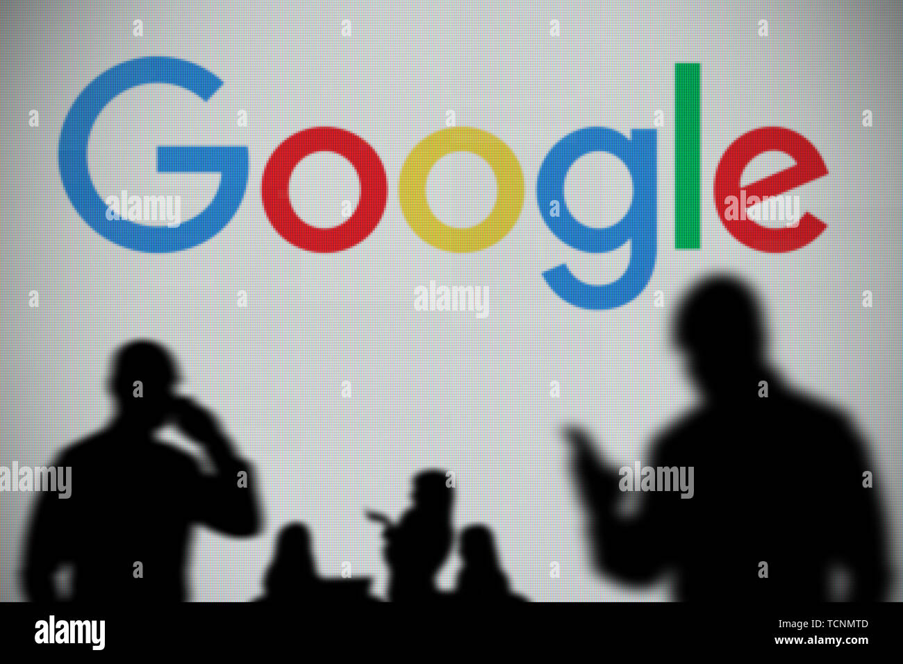 The Google logo is seen on an LED screen in the background while a silhouetted person uses a smartphone in the foreground (Editorial use only) Stock Photo