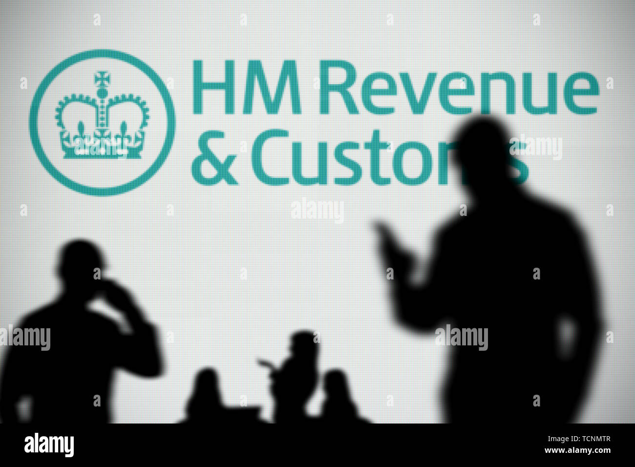 The HMRC logo is seen on an LED screen in the background while a silhouetted person uses a smartphone in the foreground (Editorial use only) Stock Photo