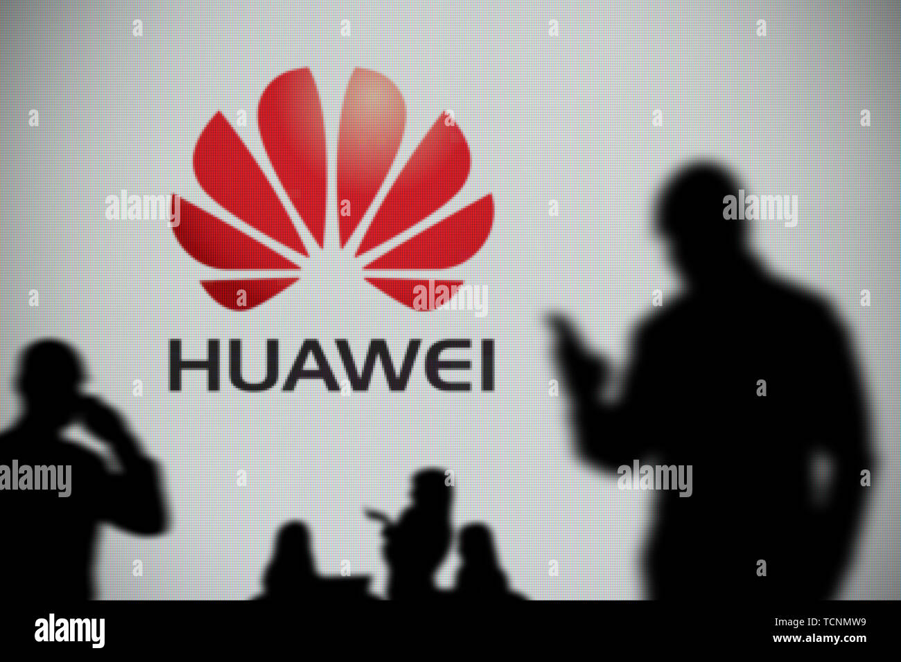The Huawei logo is seen on an LED screen in the background while a silhouetted person uses a smartphone in the foreground (Editorial use only) Stock Photo