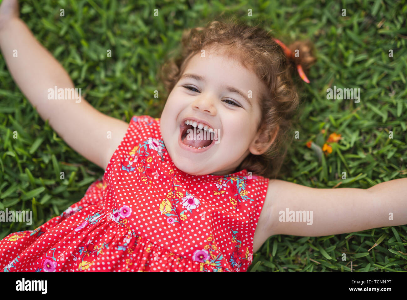 Happy Smiling Little Toddler Girl Laying On Grass In Park With Red Dress. Stock Photo