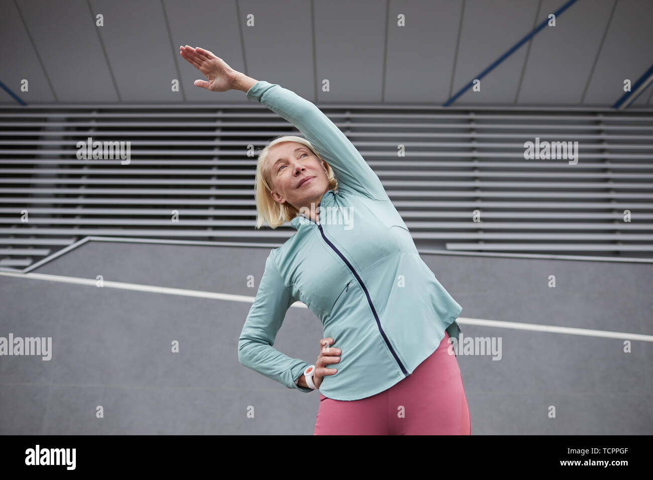 Mature active female in sportswear doing side-bends under bridge construction while spending time in urban environment Stock Photo