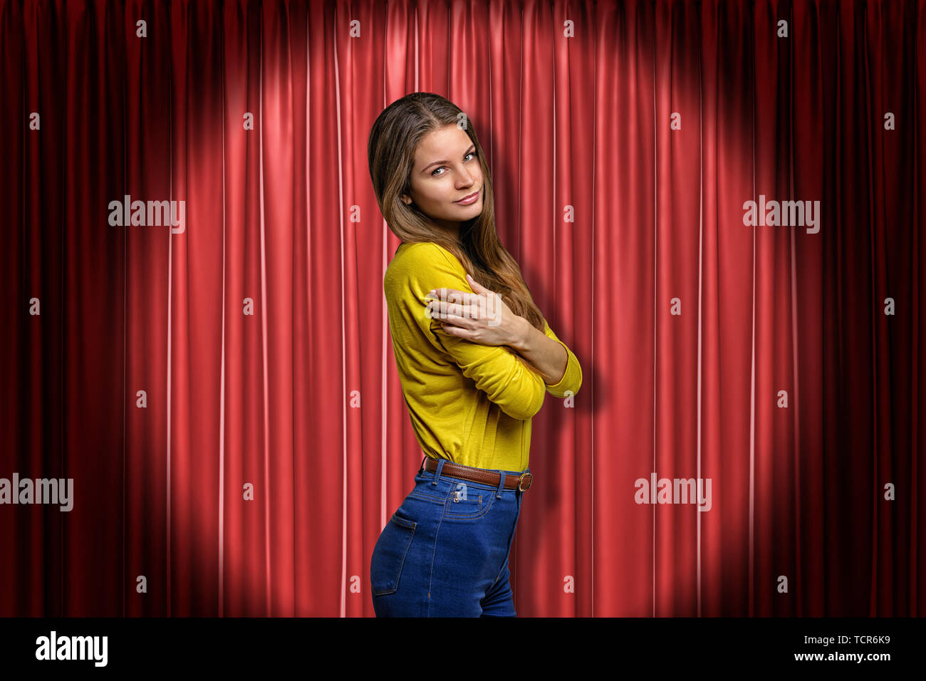 Young woman wearing blue jeans and yellow shirt embracing herself on red stage curtains background Stock Photo