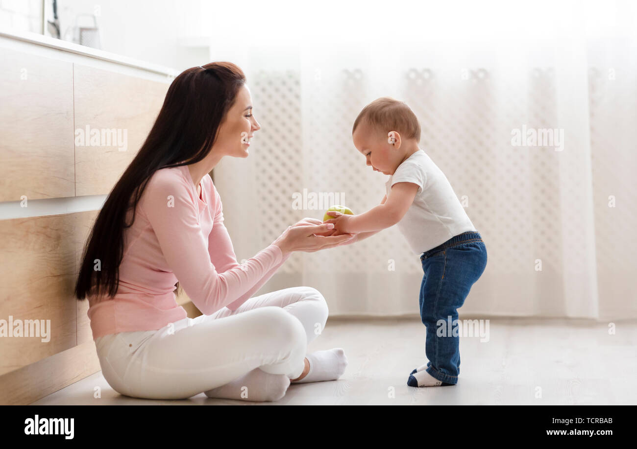 Mother giving fresh apple to baby, sitting on floor in kitchen Stock Photo