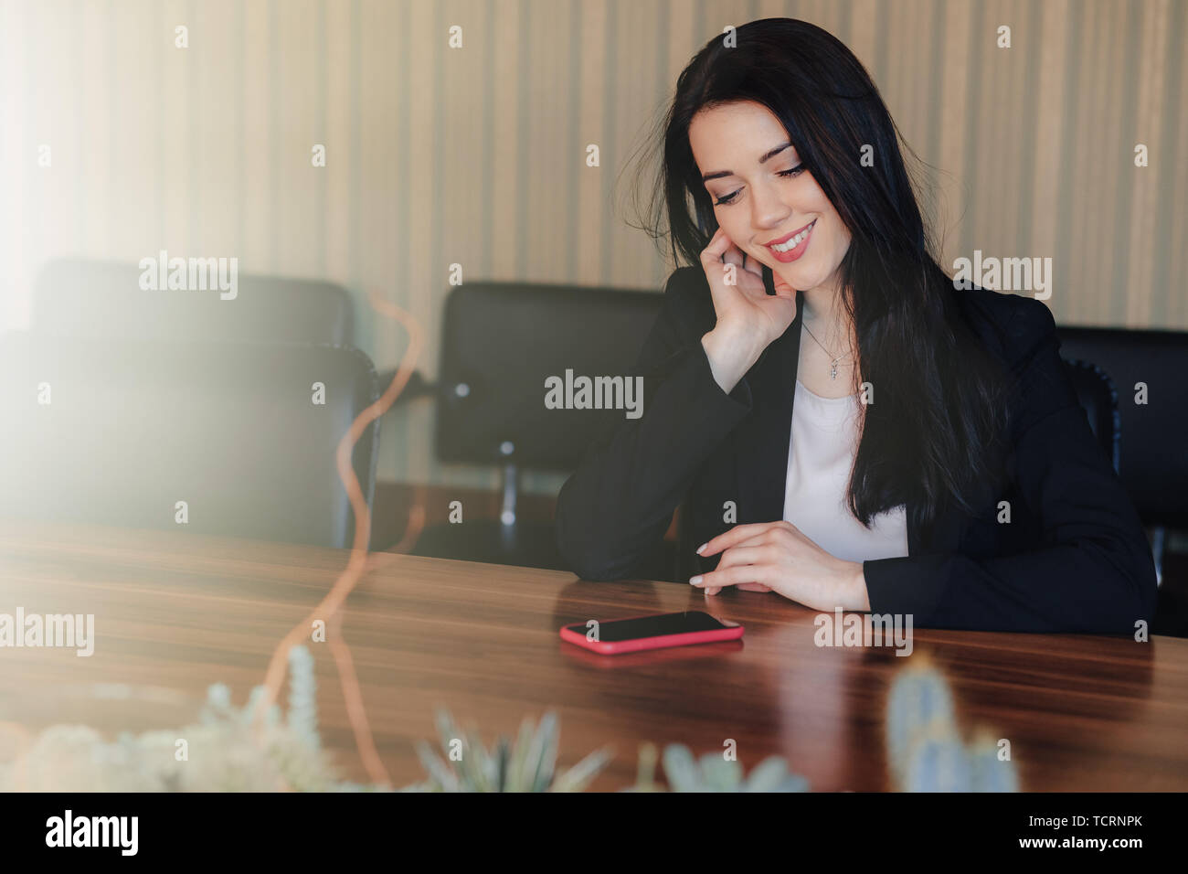Young attractive emotional girl in business style clothes sitting at desk with phone in office or audience alone Stock Photo