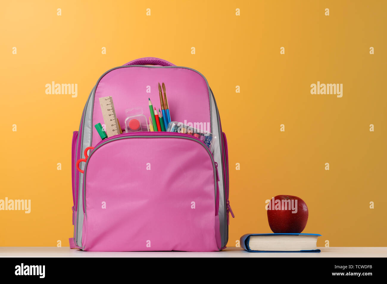 Student set. Pink backpack with office supplies, a book, a red apple on the table on a yellow background. Back to school. Stock Photo