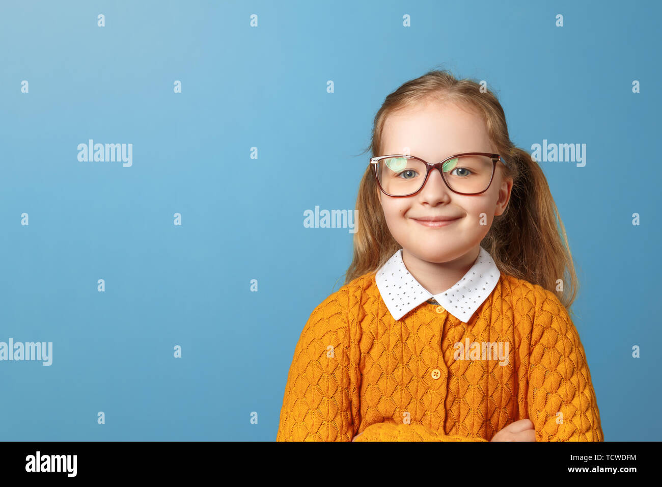 Closeup portrait of little girl schoolgirl with glasses. Pretty child in a yellow sweater on a blue background. Copy space Stock Photo
