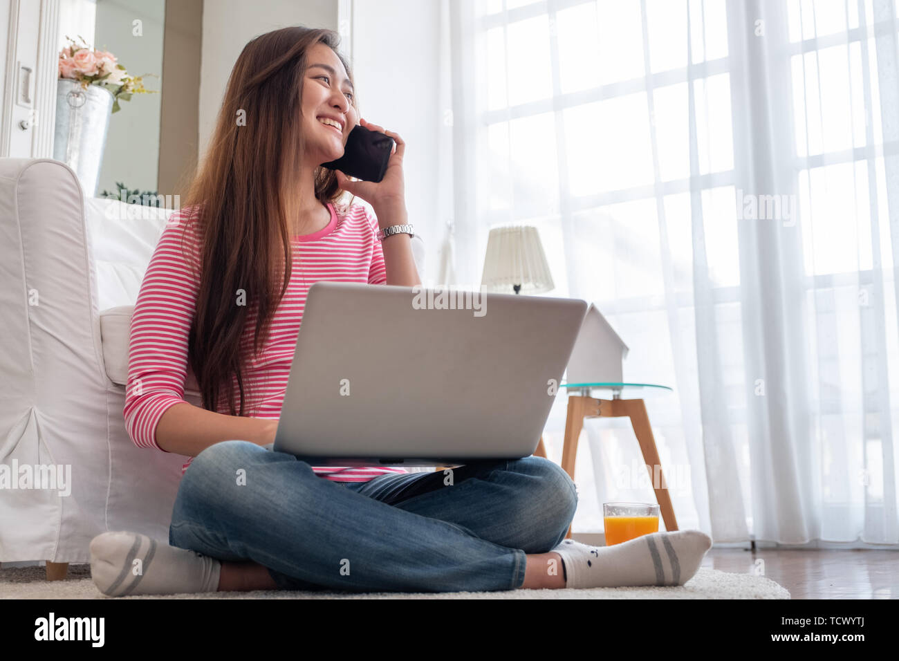 Happy asian woman talk on mobile and  typing laptop computer at home.female freelancer sitting on carpet on floor work at home in living room. Stock Photo