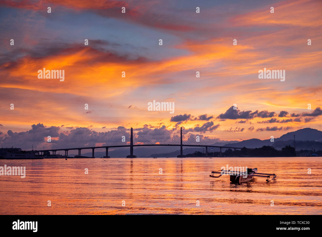 Sunset over the harbour in Ambon city showing the suspension bridge and an outrigger boat, Ambon, Moluccas (Maluku, Indonesia, Southeast Asia, Asia Stock Photo