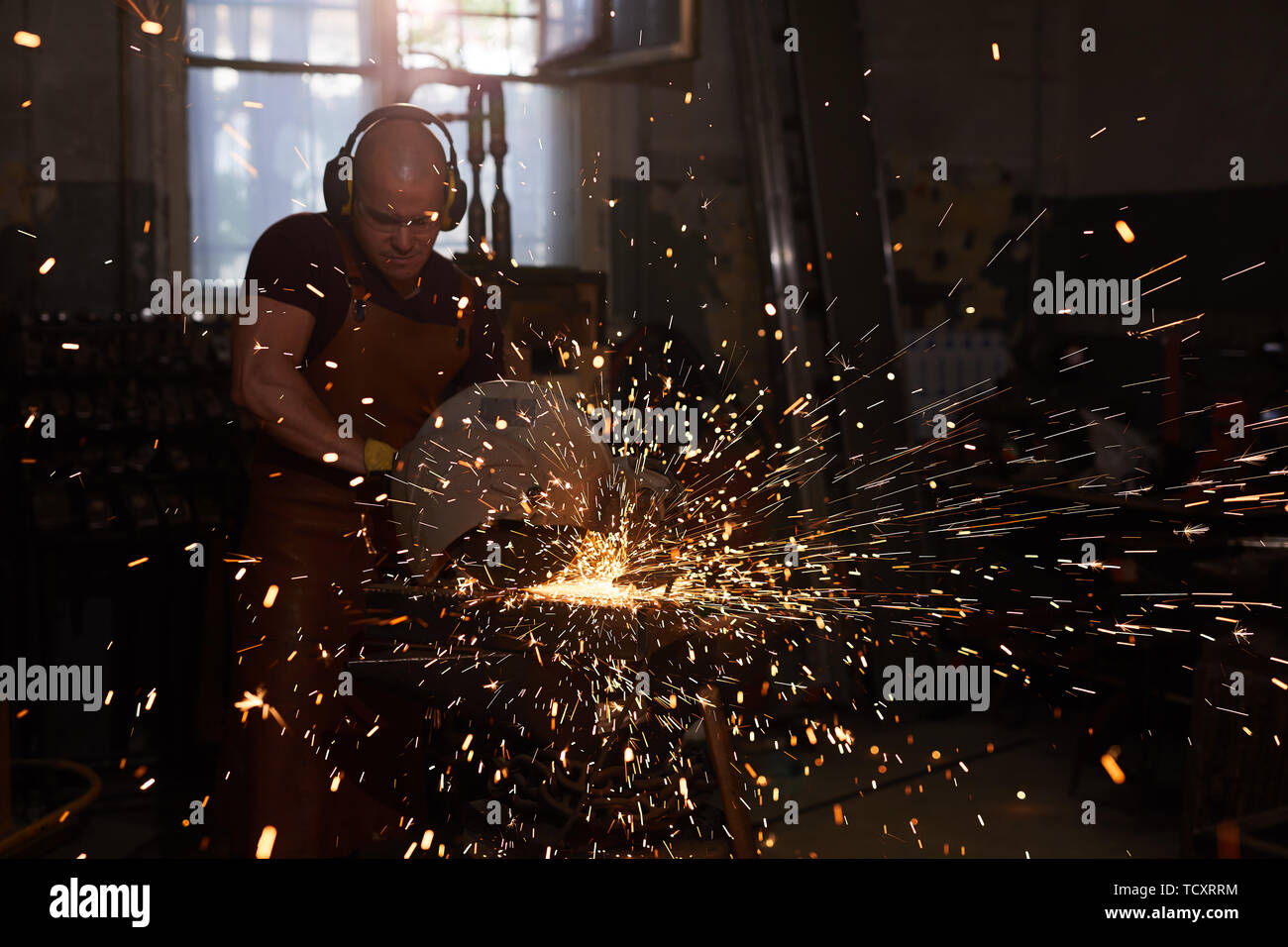 Concentrated professional blacksmith in ear protectors using cutting machine while working with metal in workshop Stock Photo