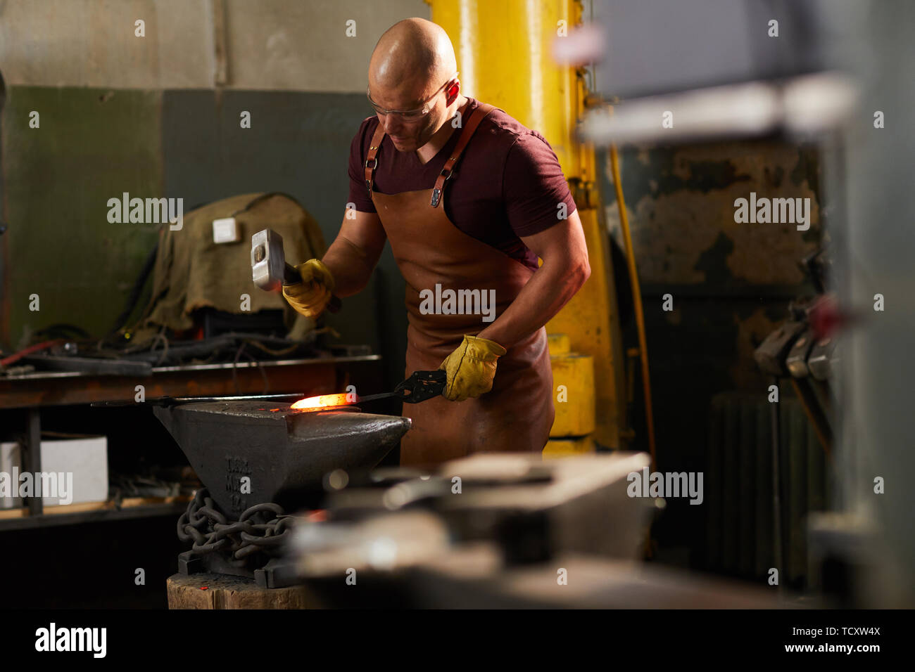 Focused young bald blacksmith in leather apron and protective goggles