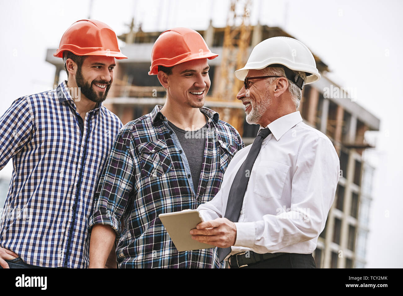 Listen carefully. Senior engineer in white helmet and formal wear is holding digital tablet and explaining building plan to young builders while stand Stock Photo