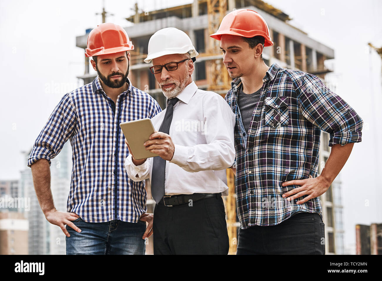 Using modern technologies. Senior engineer is looking at project plan on digital tablet with two young builders while working at construction site. Te Stock Photo