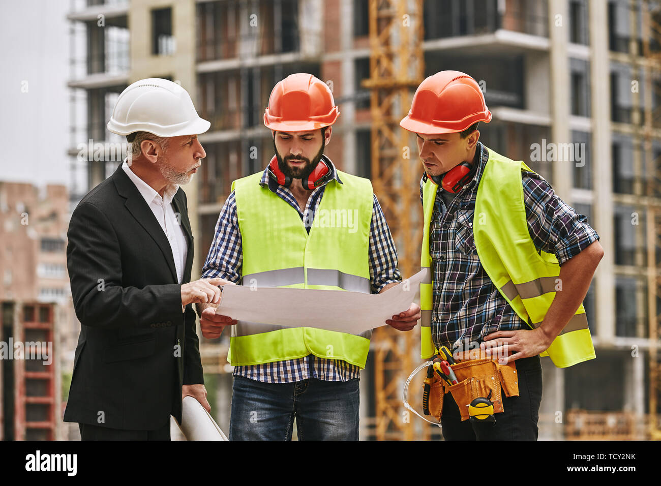 Team meeting. Main engineer in formal wear and white helmet with two young builders are discussing a construction drawing and main concept of building Stock Photo
