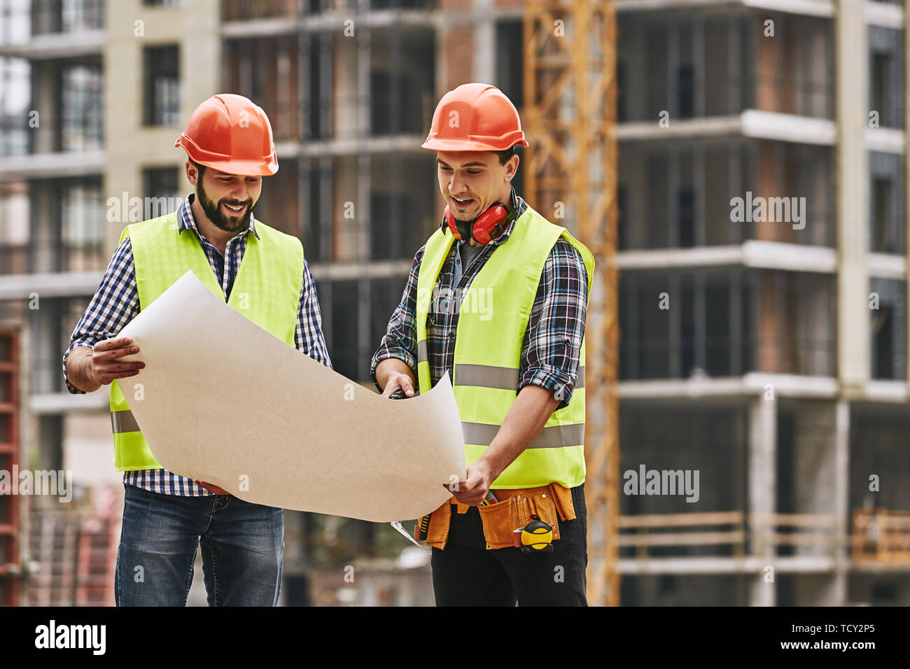 Look here! Two young builders in working uniform and helmets are holding a construction drawing in their hands and discussing it with each other while Stock Photo