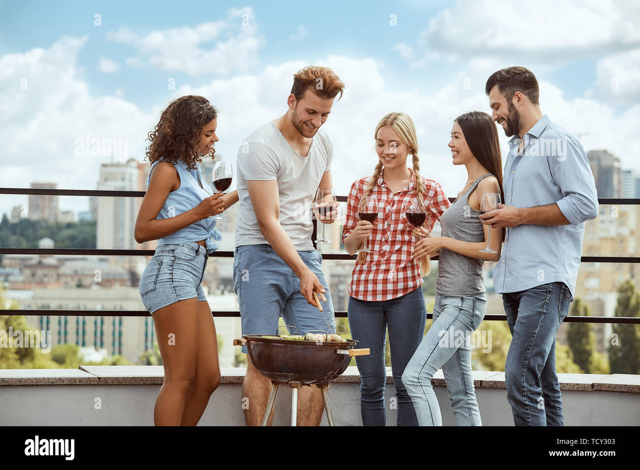 Perfect time for barbeque. Young man barbecuing meat on the grill while drinking wine and spending a time with friends. on the roof. Picnic. Summer ti Stock Photo