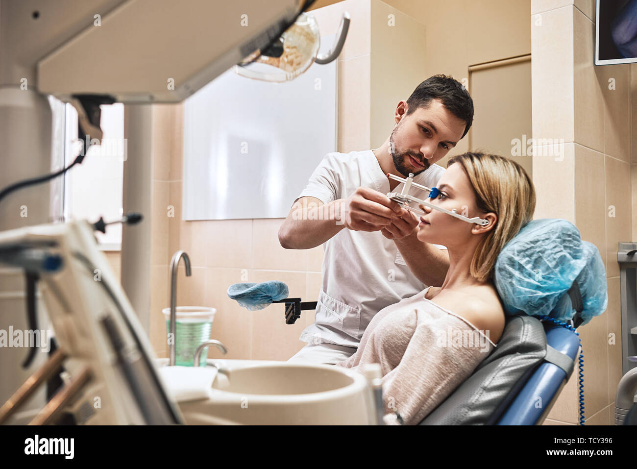 Portrait of attractive woman sitting in the dental chair and listening to her dentist. He stands near her and checks the symmetry of her jaws using or Stock Photo