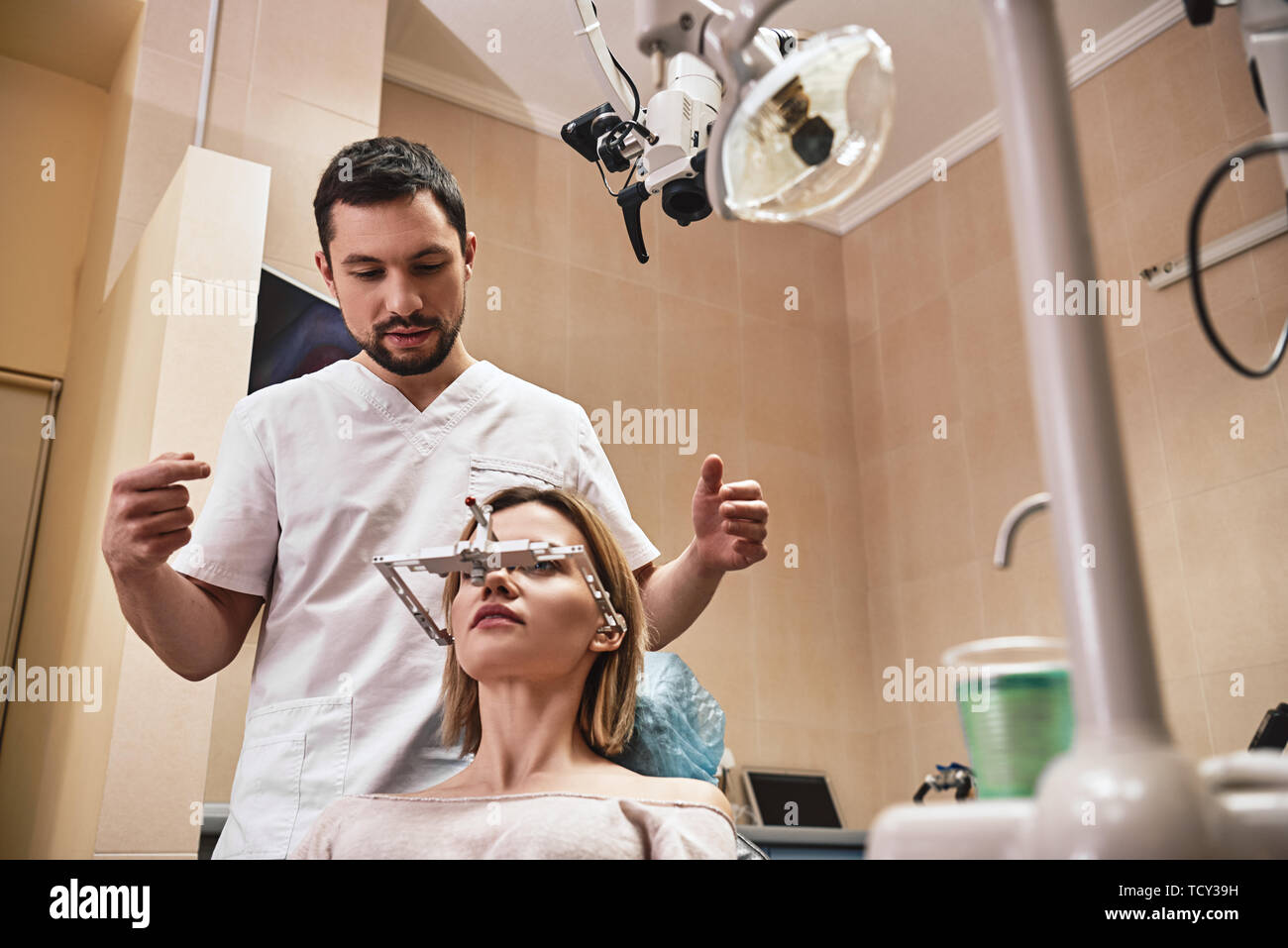 Portrait of attractive woman sitting in the dental chair and listening to her dentist. He stands near her and checks the symmetry of her jaws using or Stock Photo