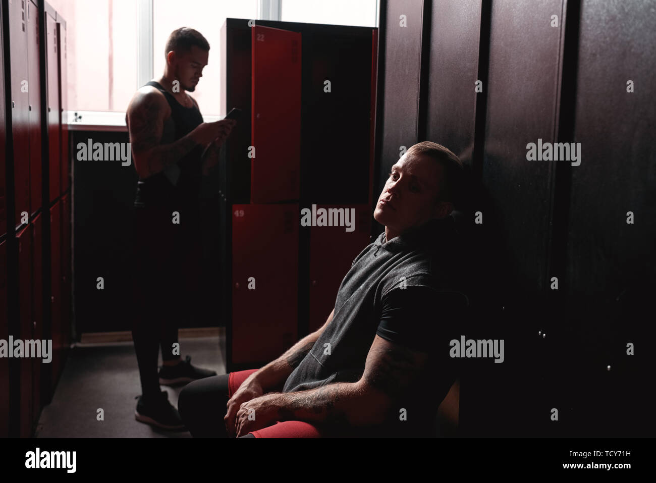 Thoughtful young muscular athlete in sports clothing waiting for training while sitting in dark gym locker room Stock Photo