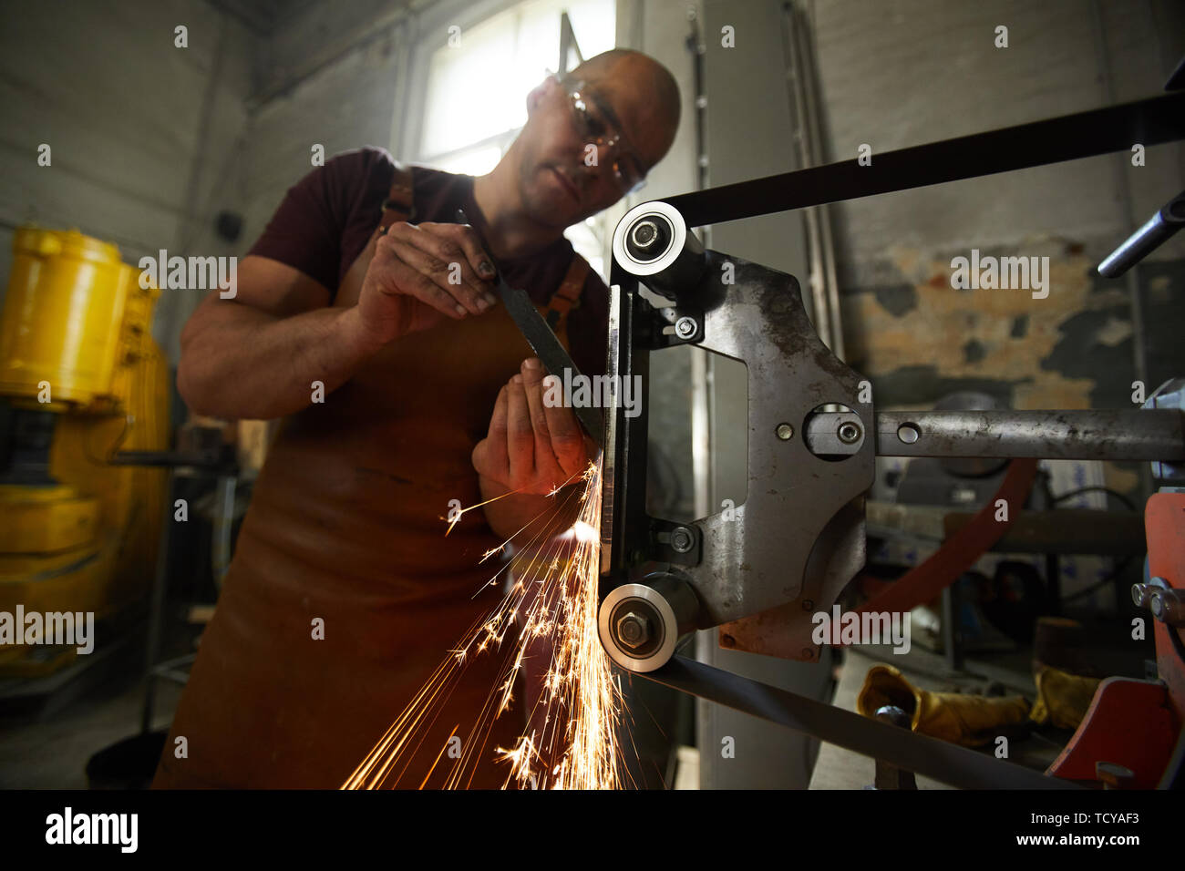 Concentrated young blacksmith in apron standing at grinding machine and
