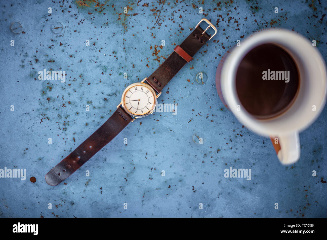 Vintage wrist watch on rustic blue metal bench suggesting the time is 07.00, a cup of coffee in the edge of frame. Stock Photo