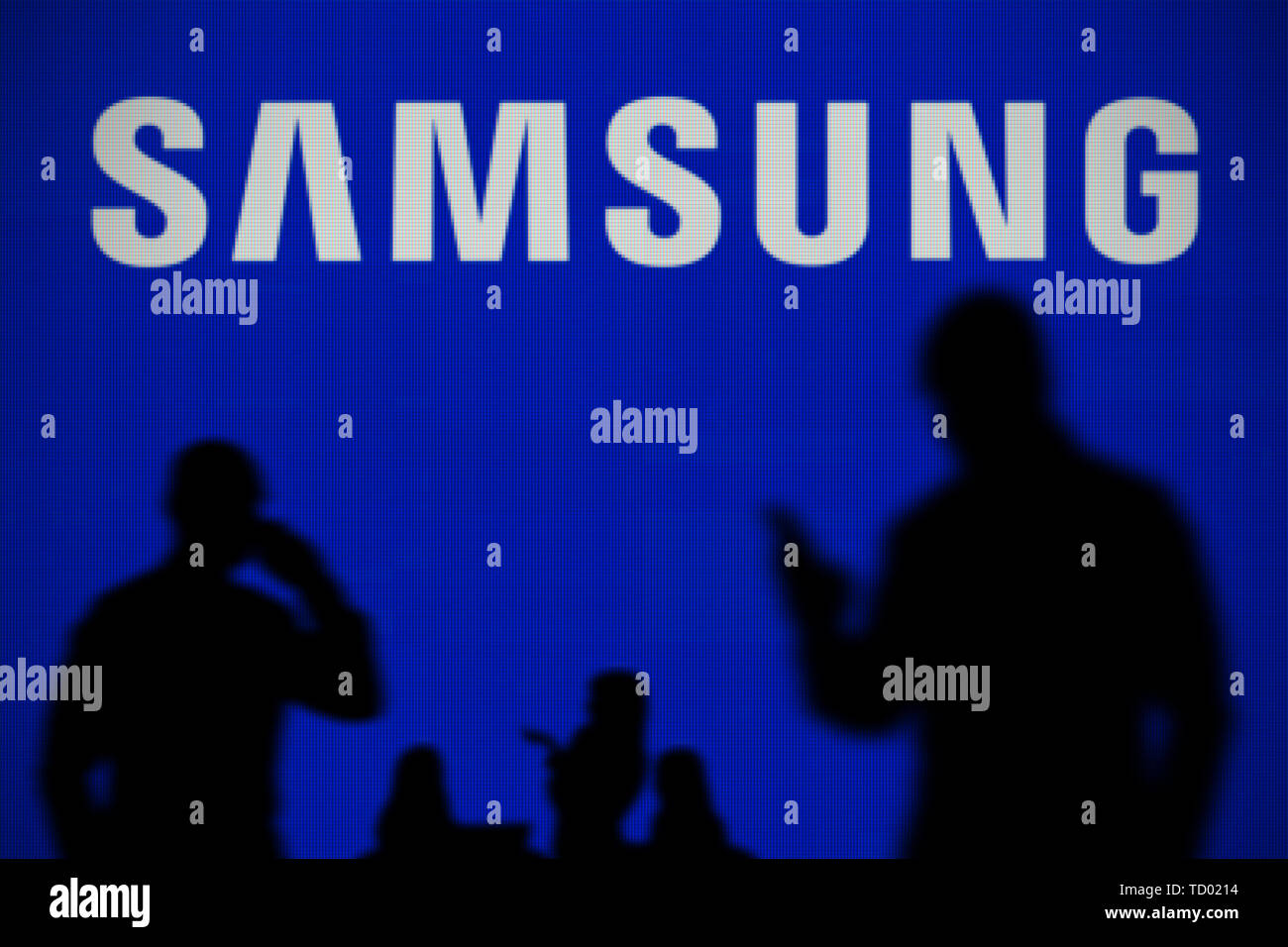 The Samsung logo is seen on an LED screen in the background while a silhouetted person uses a smartphone in the foreground (Editorial use only) Stock Photo