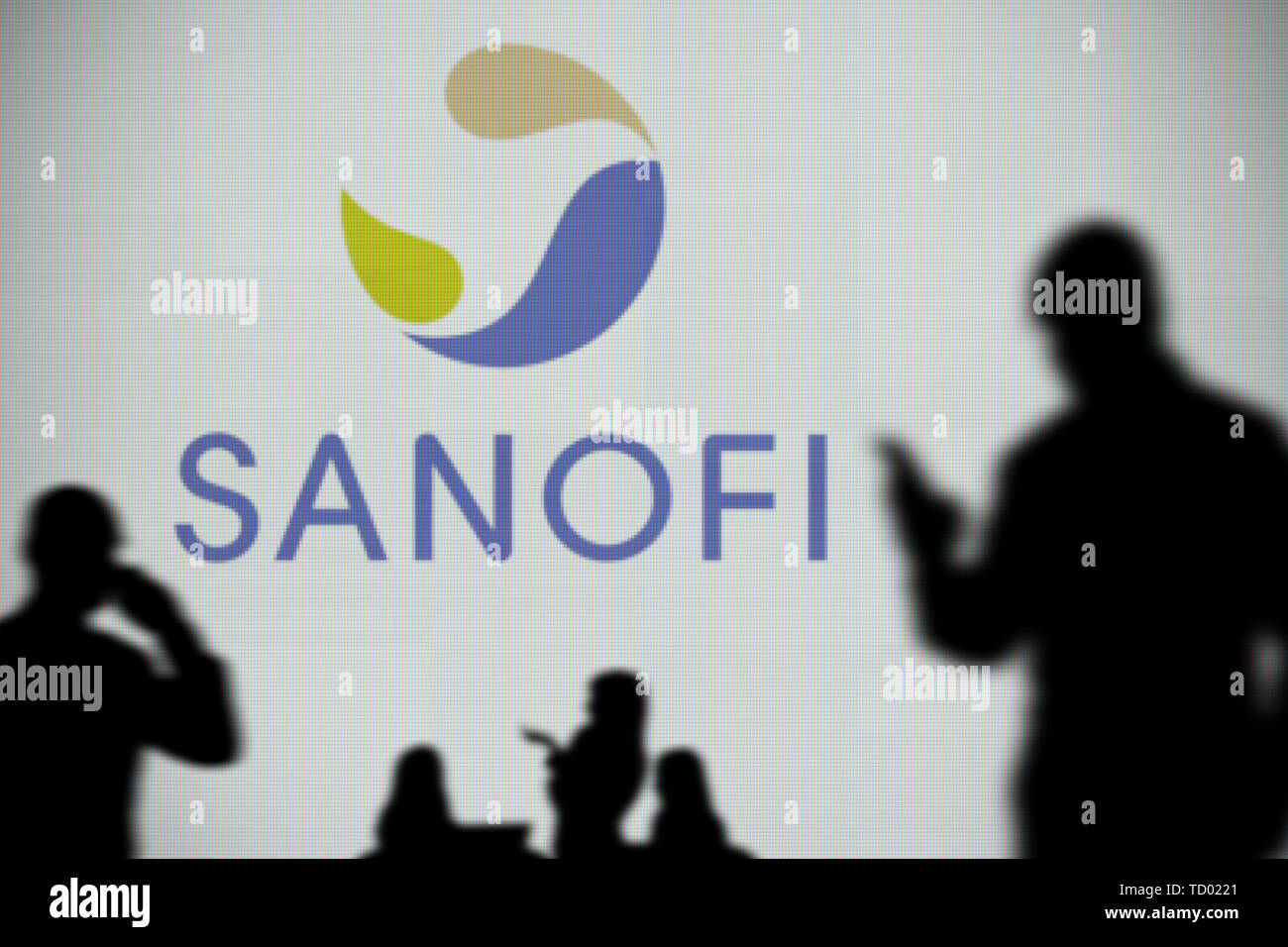The Sanofi logo is seen on an LED screen in the background while a silhouetted person uses a smartphone in the foreground (Editorial use only) Stock Photo