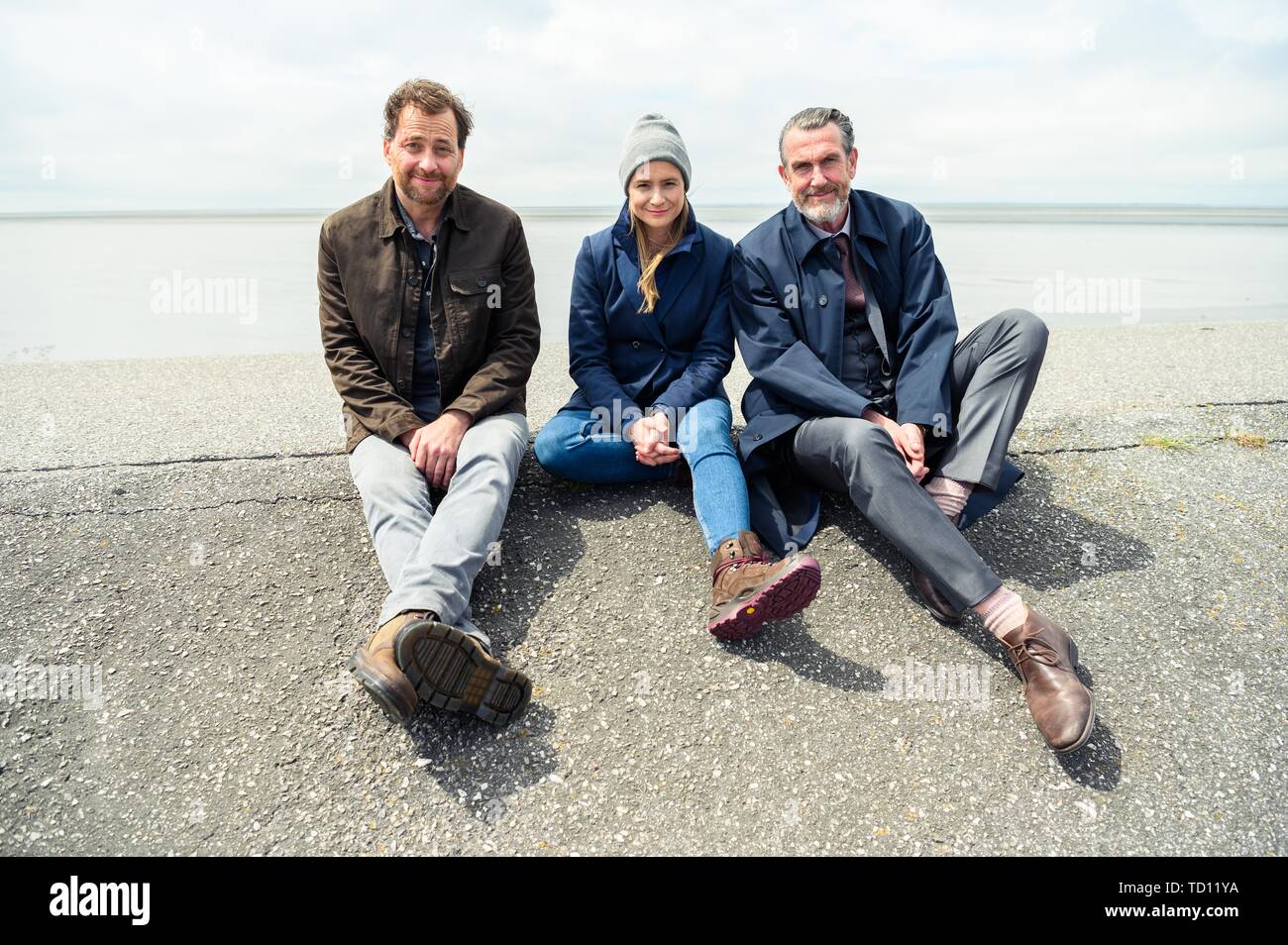 Norddeich, Germany. 11th June, 2019. Christian Erdmann (as Frank Eller; l-r), Julia Jentsch (as Ann Kathrin Klaasen), and Kai Maertens (as Ubbo Heide) sit on the dike during the press conference for the Saturday thriller 'Ostfriesengrab'. The fourth film in the Ostfriesland crime series is based on the novel of the same name by Klaus-Peter Wolf (about dpa 'successful author Klaus-Peter Wolf provides material for new ZDF crime thriller'). Credit: Mohssen Assanimoghaddam/dpa/Alamy Live News Stock Photo