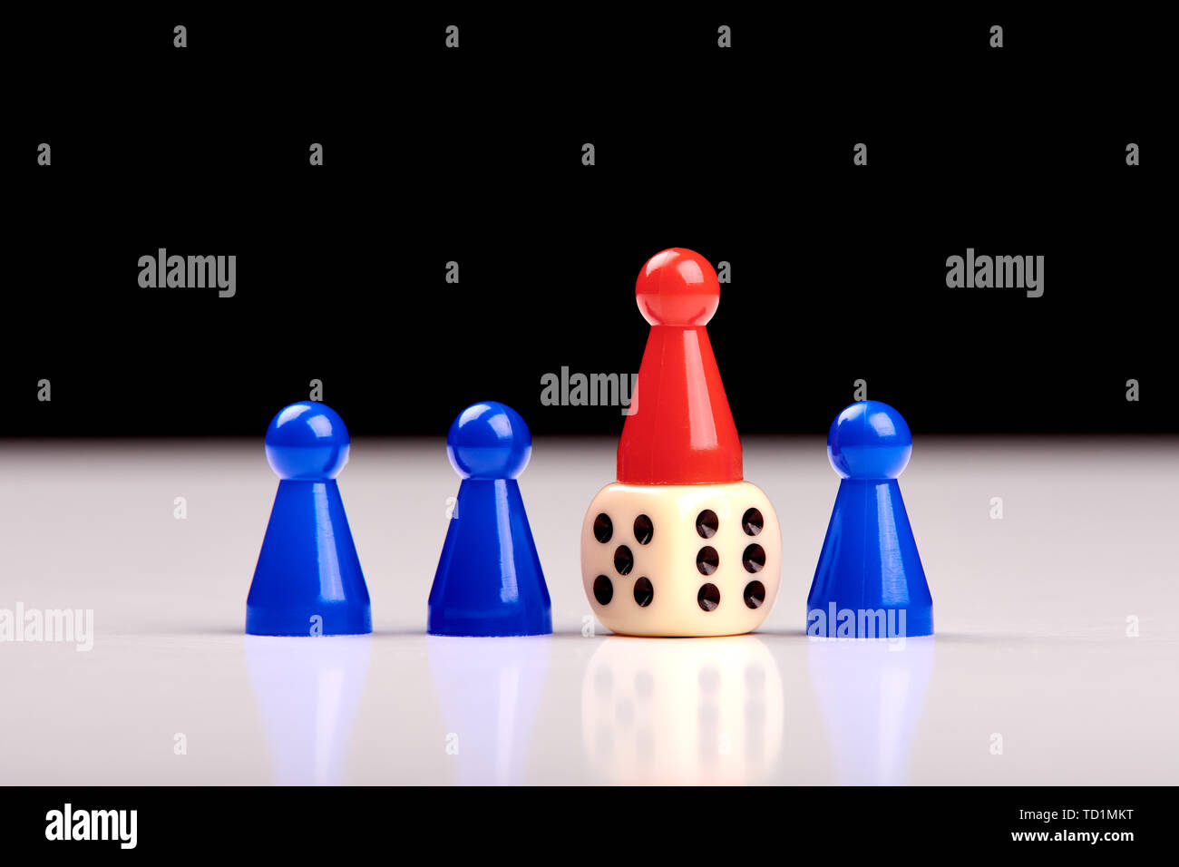 Three blue game pieces and between them one red piece stands on a dice as a winner or leader. Blurred black and white background Stock Photo