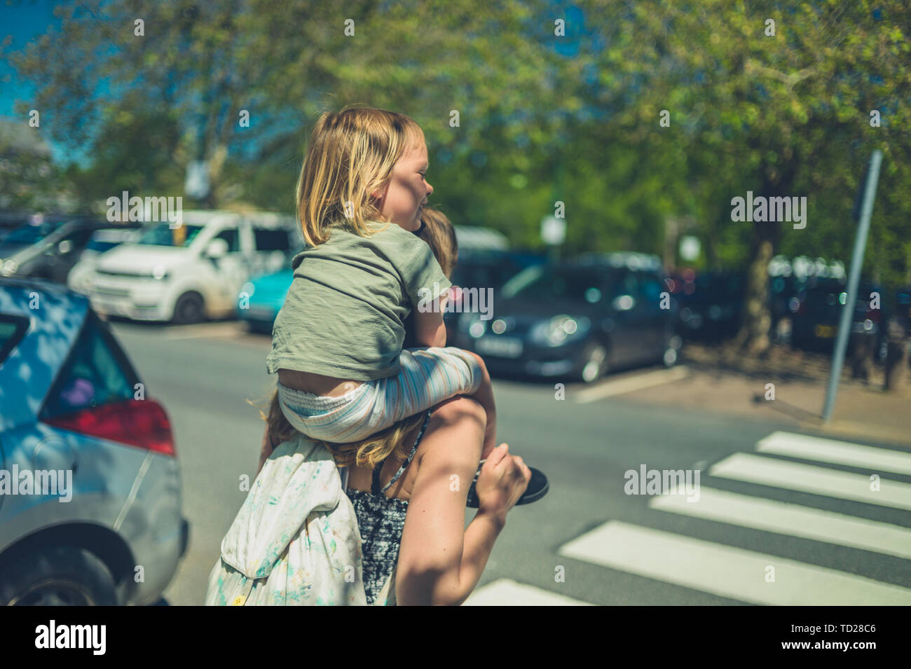 A little toddler is riding on his mother's shoulders in a car park on a sunnny summer day Stock Photo