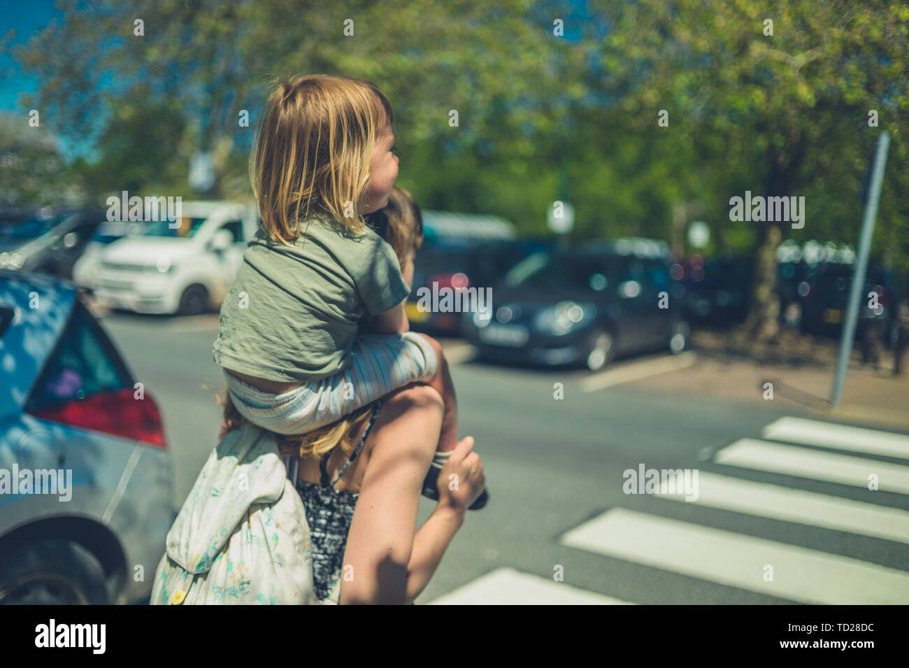 A little toddler is riding on his mother's shoulders in a car park on a sunnny summer day Stock Photo