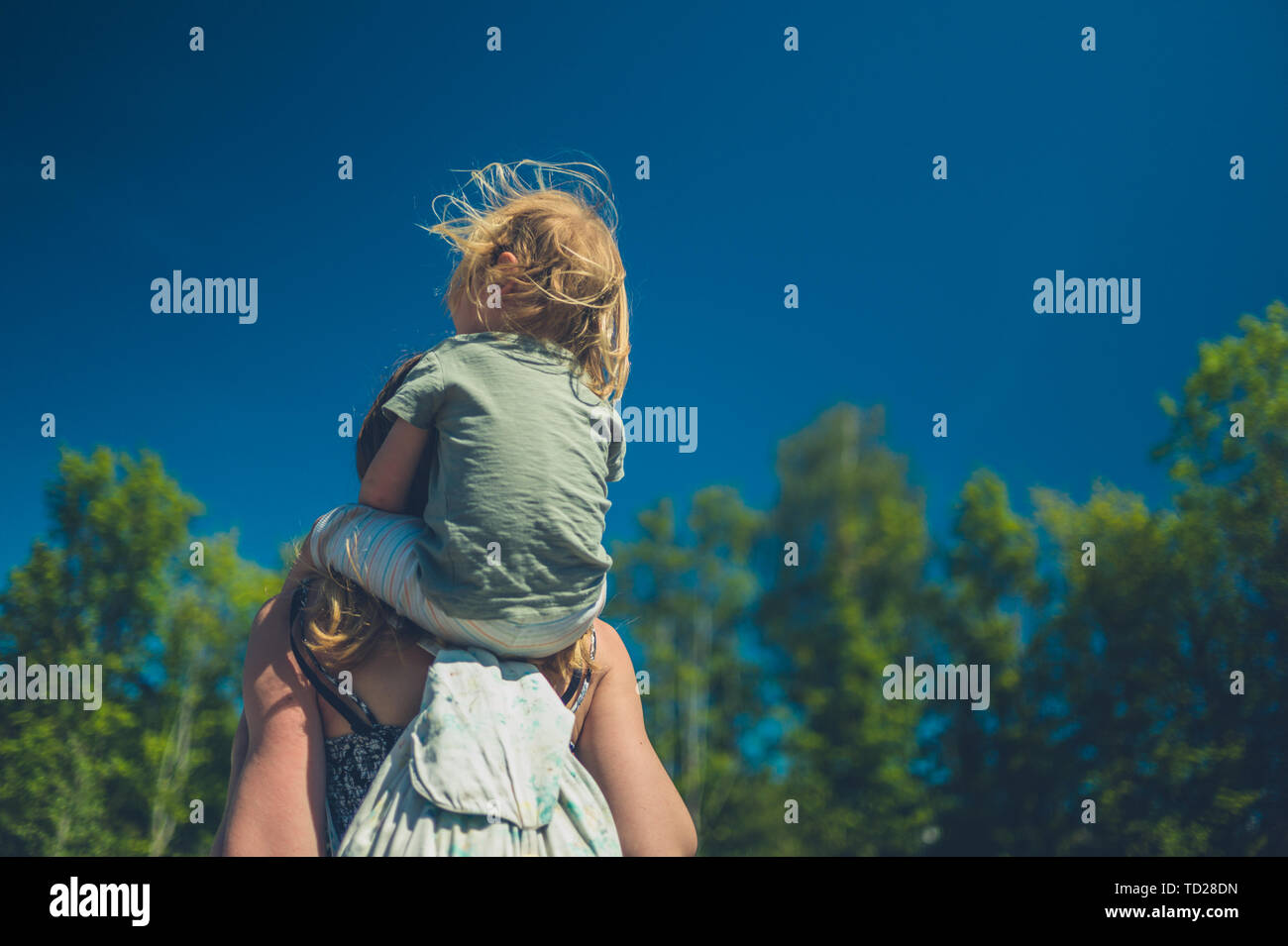 A little toddler is riding on his mother's shoulders outdoors on a sunny summer day Stock Photo
