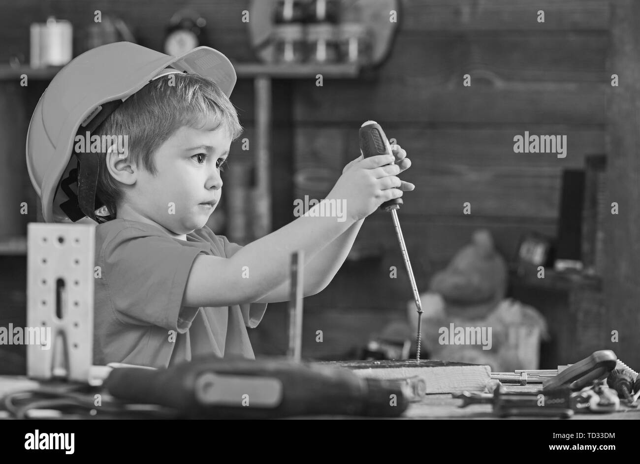 Side view little kid binding screw into wooden block. Concentrated boy in orange helmet sitting at table and holding screwdriver. Stock Photo