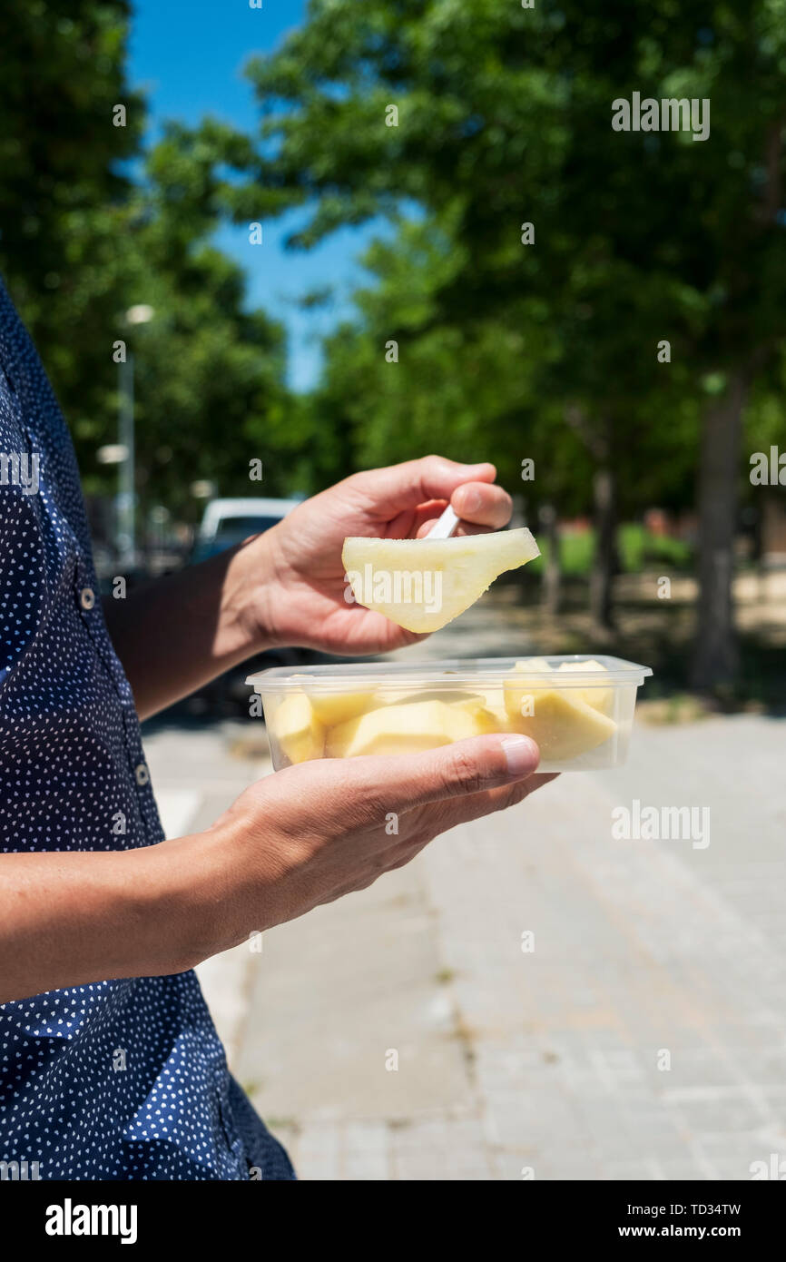 closeup of a young caucasian man outdoors, wearing a blue short-sleeved shirt, eating a salad fruit, made with pear, apple and peach, from a plastic c Stock Photo