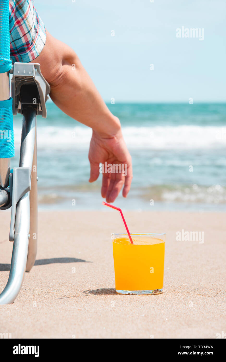 closeup of a young caucasian man relaxing on the beach, sitting on a deck chair next to a glass with a refreshing orange drink placed on the sand, wit Stock Photo