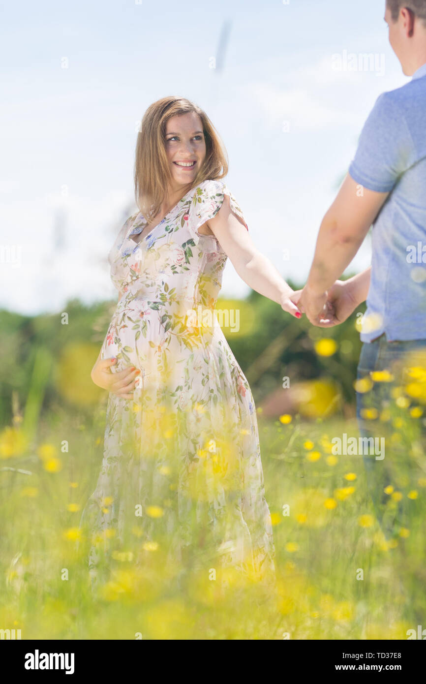 Young happy pregnant couple in love holding hands, relaxing in meadow. Stock Photo