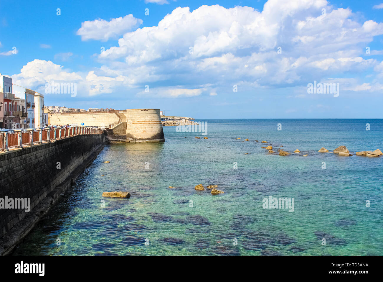 Amazing view of blue sea and adjacent city coast in Syracuse, Sicily, Italy on a sunny day with blue sky. Stones in sea water. Photographed in Ortygia Island. Popular tourist place. Stock Photo