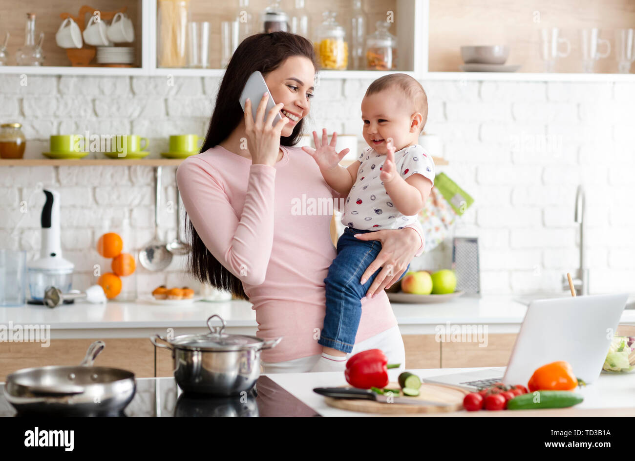Mother talking on smartphone and holding adorable baby in kitchen Stock Photo