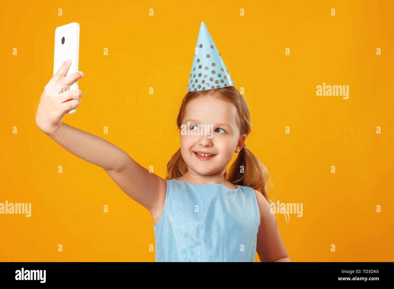 Cheerful little girl celebrates birthday. The child holds the phone, takes a selfie. Closeup portrait on yellow background. Stock Photo