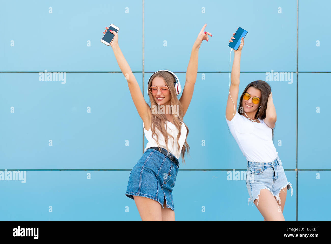 Fashion portrait of two pretty smiling hipsters woman in sunglasses holding smartphone and dancing against the colorful blue wall Stock Photo