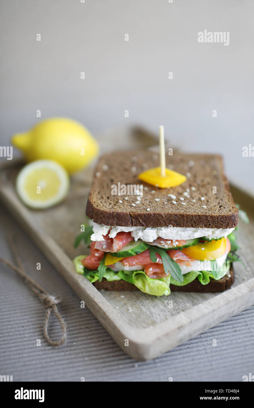 Sandwich with smoked salmon red fish, cheese, green salad, cucumber, arugola, sweet pepper and sesame seeds on rye bread in wooden plate. Stock Photo