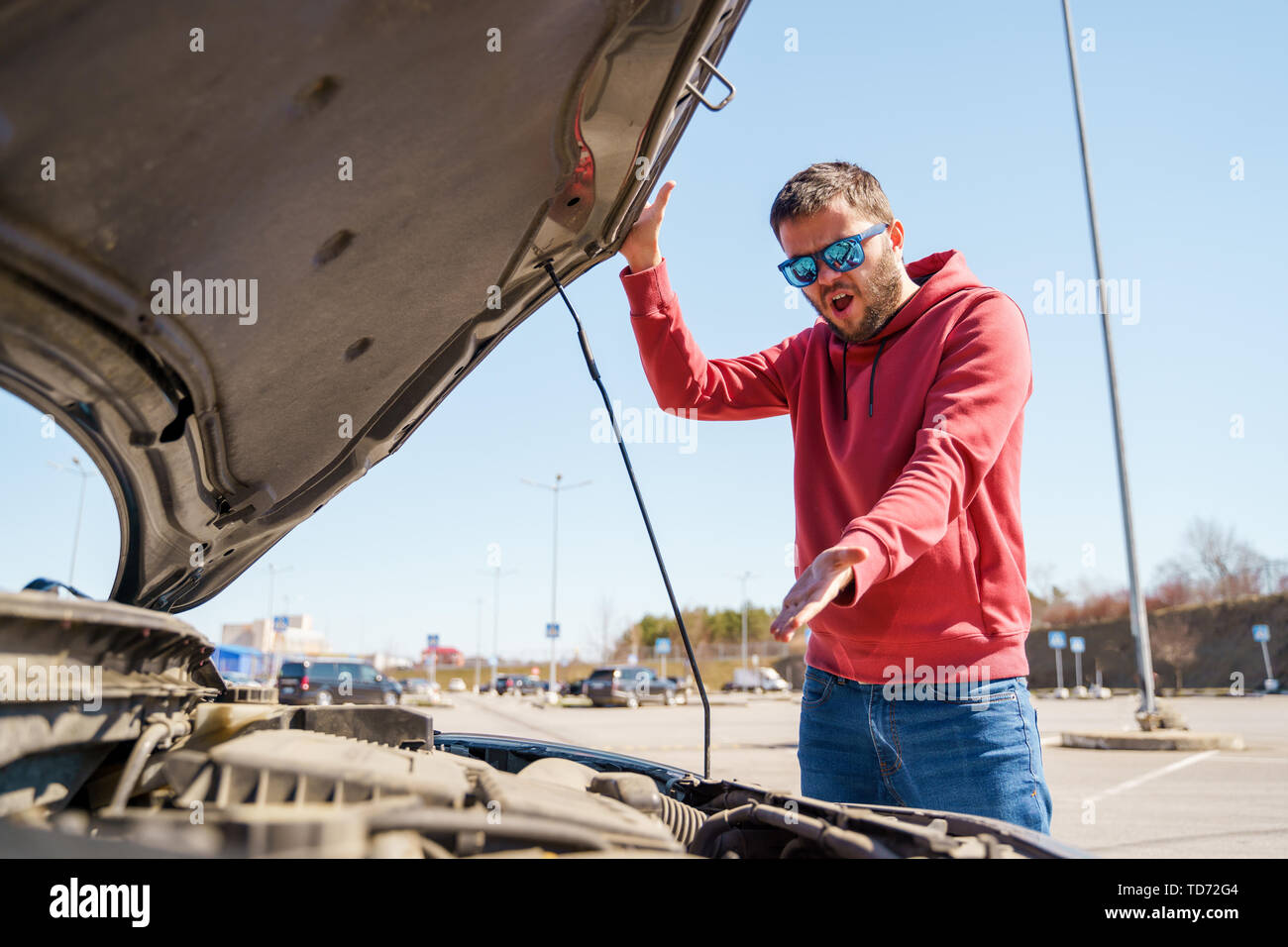 Photo of upset man next to open hood of broken car in daytime Stock