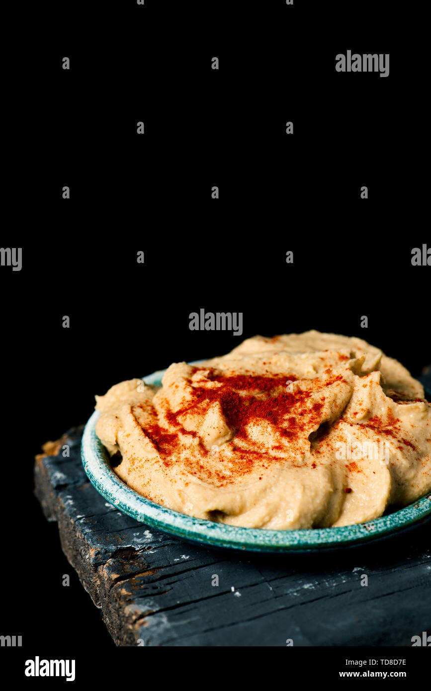 a homemade hummus seasoned with paprika served in a green ceramic plate, on a dark gray rustic wooden table against a black background with some blank Stock Photo