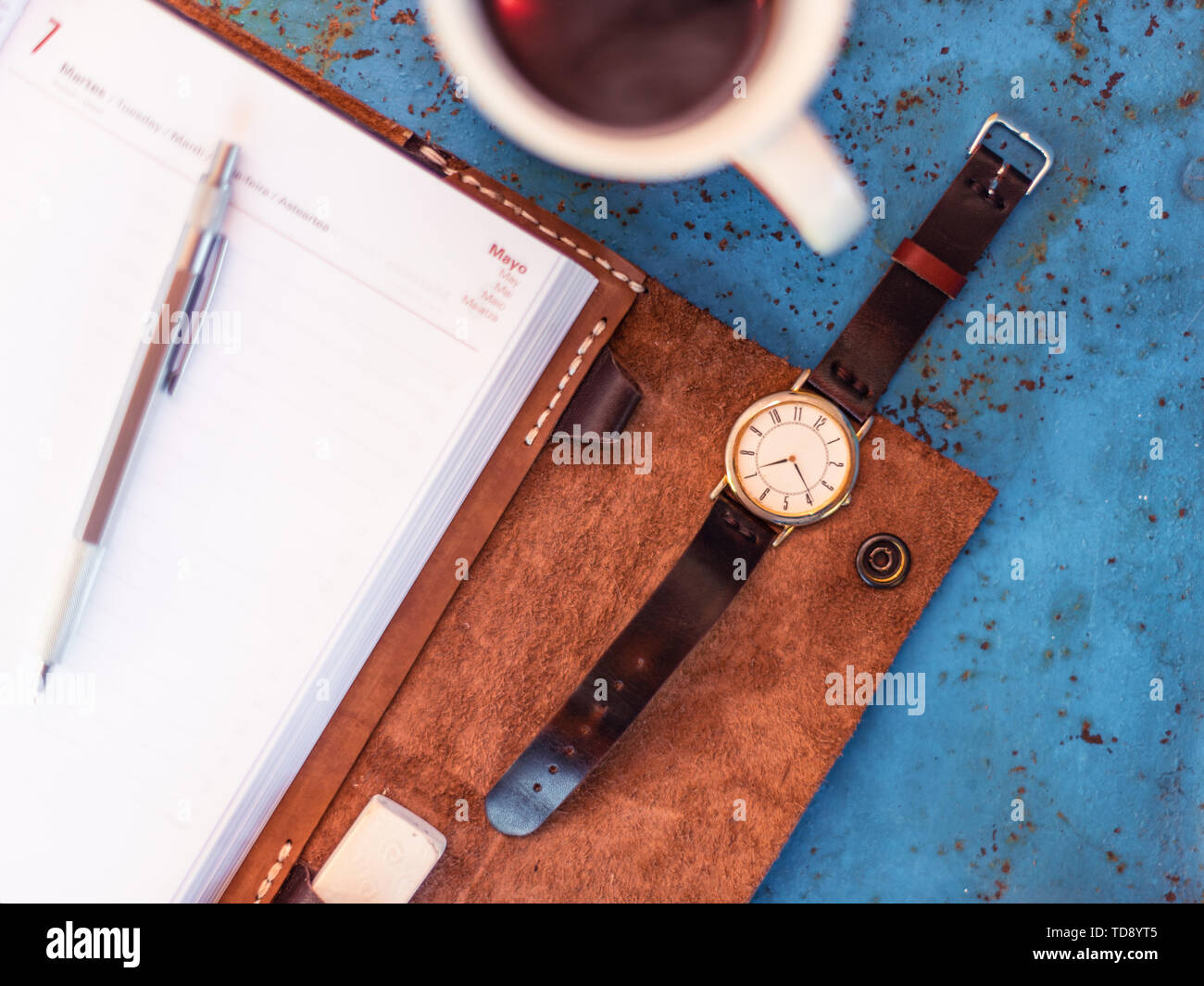 Vintage wrist watch and leather diary on rustic blue metal bench suggesting the time is 07.00, a cup of coffee in the edge of frame. Stock Photo