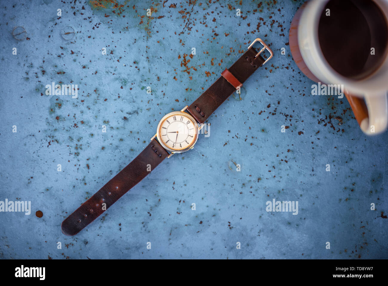 Vintage wrist watch on rustic blue metal bench suggesting the time is 07.00, a cup of coffee in the edge of frame. Stock Photo