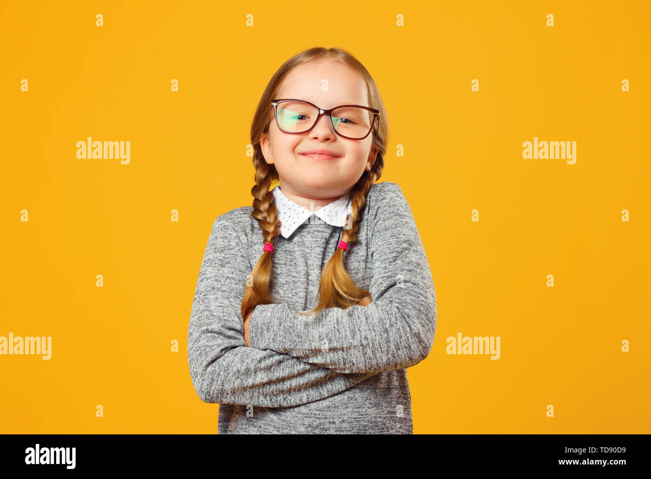 Closeup portrait of a cheerful little girl in glasses on yellow background. Child schoolgirl folded her arms and looks into the camera. Stock Photo