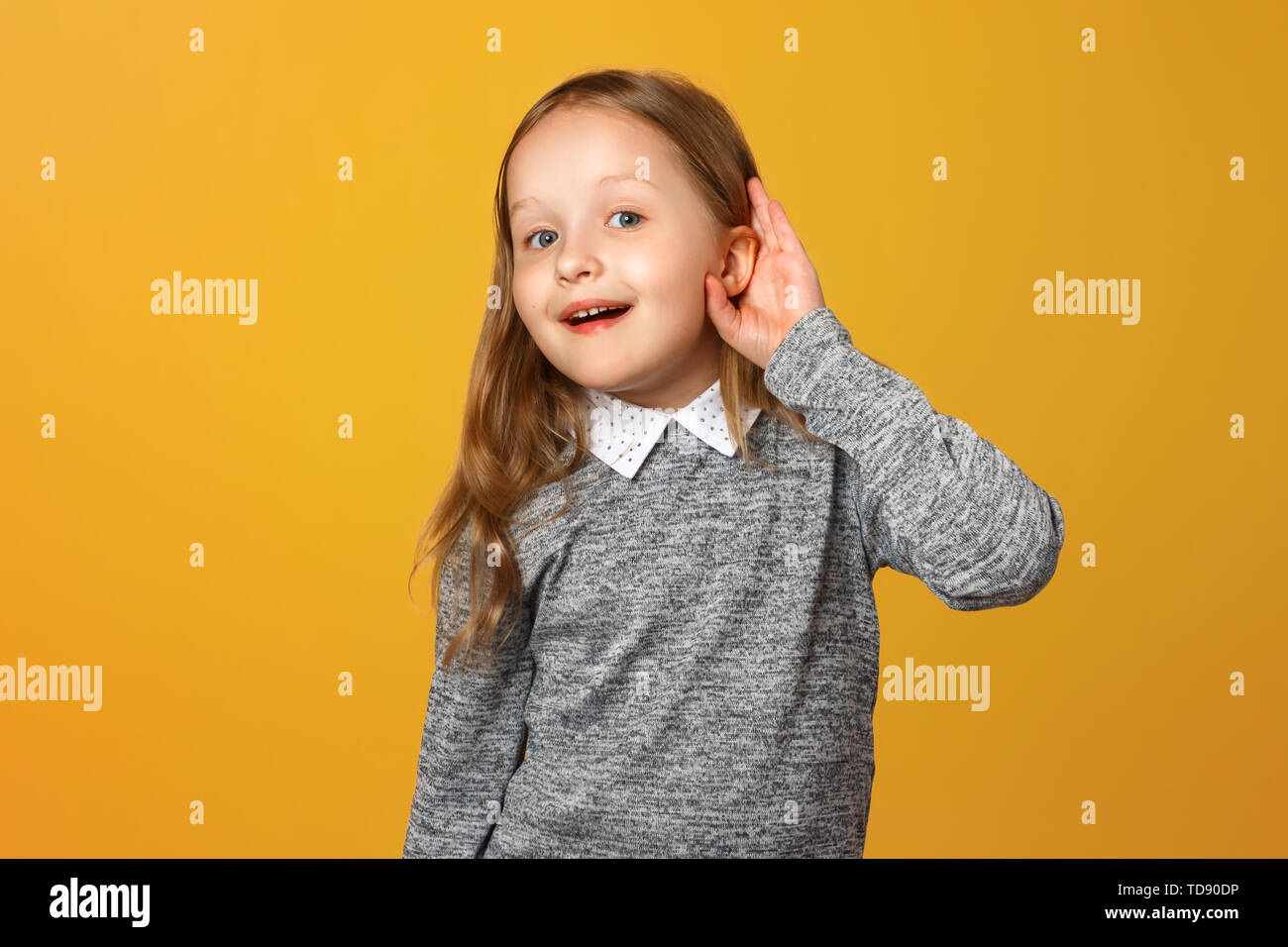 The little girl puts her hand to her ear to hear better. The child is listening to something on a yellow background. Stock Photo